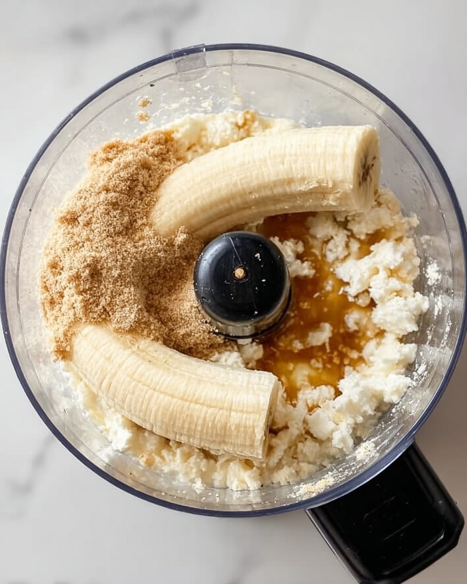 The image shows a food processor from above filled with several layers of ingredients. The bottom layer is white cottage cheese with small chunks, showing a soft texture. On top of it, there are two whole peeled bananas placed lengthwise on opposite sides. Covering some of the bananas and cottage cheese is a powdery light brown layer, likely a type of flour or crushed oats. A small amount of amber syrup is drizzled near the center, creating a shiny contrast. The food processor is on a white marbled surface. Photo taken with an iphone --ar 4:5 --v 7