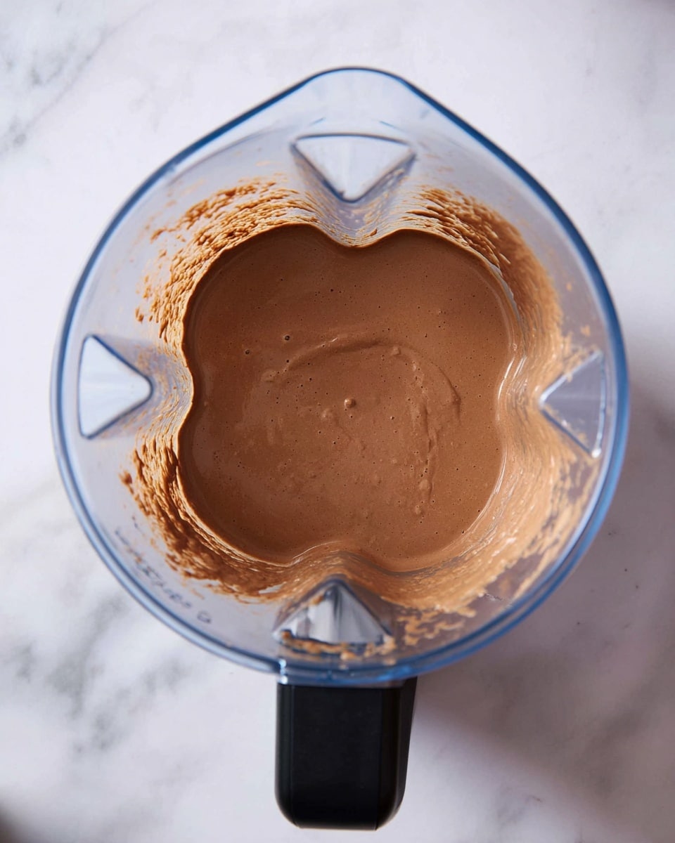 A clear blender pitcher is shown from above, filled with a smooth, thick, light brown chocolate mixture that fills about half the pitcher. The mixture has a velvety texture with no lumps, and some of it clings to the sides of the pitcher near the blades at the bottom. The blender pitcher has a black handle on the left, and it is placed on a white marbled surface. photo taken with an iphone --ar 4:5 --v 7