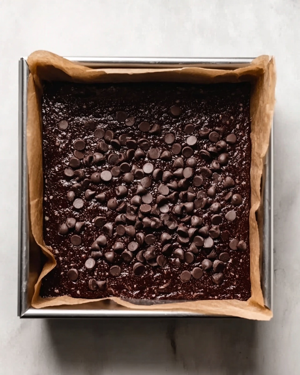 The image shows a square metal baking pan lined with parchment paper, filled with a thick dark brown batter. On top of the batter are scattered dark brown chocolate chips that look slightly shiny and smooth. The pan sits on a white marbled surface, and the batter appears dense and rich, covering the entire base of the pan evenly. Photo taken with an iphone --ar 4:5 --v 7