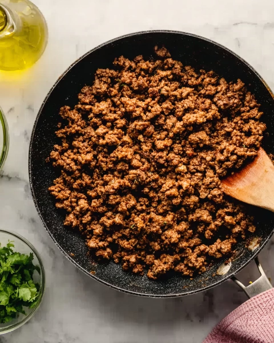 A close-up view of a black pan filled with cooked ground meat, showing crumbled pieces with a browned and slightly crispy texture. A wooden spatula is partially visible in the pan, with a woman's hand holding its handle from the right side. The pan is on a white marbled surface, next to a small bowl with fresh green herbs and a clear glass bottle containing light yellow oil. The lighting highlights the rich color of the cooked meat, making it appear juicy and well-seasoned. Photo taken with an iphone --ar 4:5 --v 7