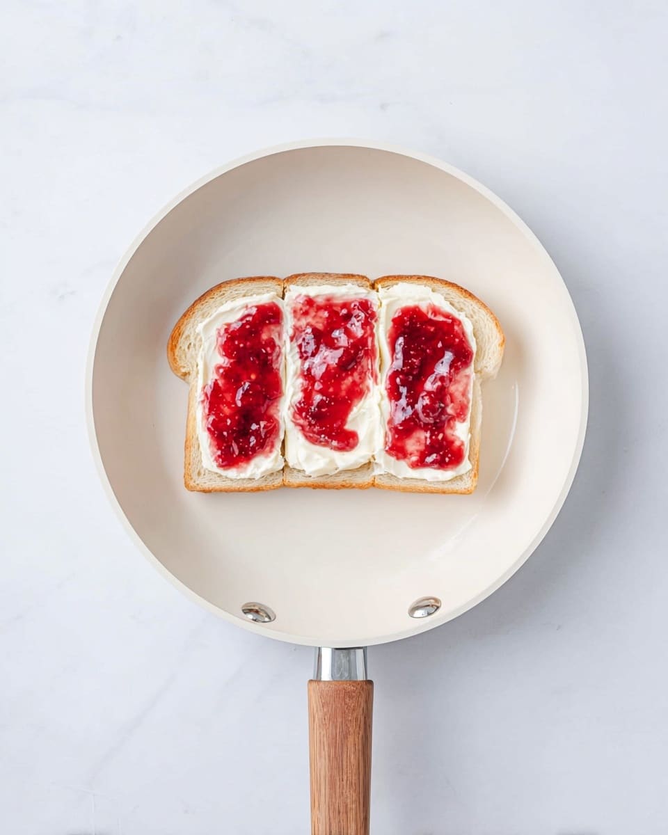 A slice of white bread is placed in the center of a white frying pan with a wooden handle. The bread has three slices of soft cream cheese evenly spread on top, forming the first layer. Over this, a bright red jam with visible fruit pieces is spread thickly in three sections, covering the cream cheese but not the bread edges. The white frying pan rests on a white marbled surface, showing a clean and bright setting. Photo taken with an iphone --ar 4:5 --v 7
