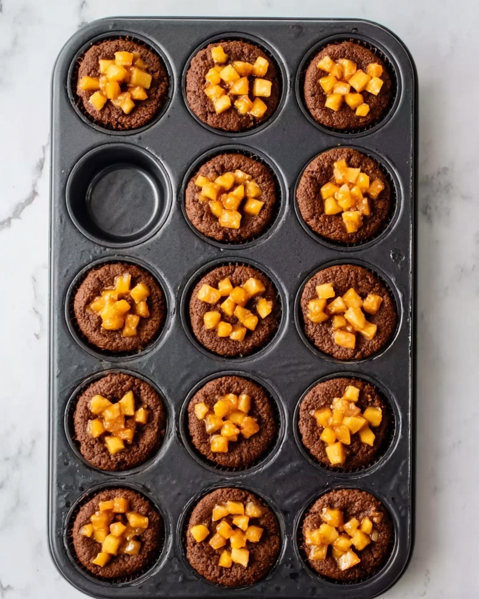 A black muffin tray holds 16 baked muffins with a deep brown outer layer and a slightly cracked surface. Each muffin is topped with a heap of small orange and yellow diced fruit pieces resembling cooked apples or peaches, placed in the center as a bright, textured layer. Four empty muffin slots are on the left side of the tray. The tray rests on a white marbled surface with light gray veining visible around it. photo taken with an iphone --ar 4:5 --v 7