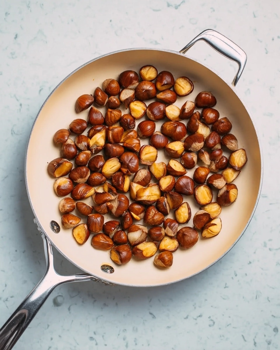 The image shows a white frying pan filled with small, evenly browned chestnut pieces, spread across the pan’s surface. The chestnuts vary in shades of golden brown to dark brown, with a slight crispy texture visible. The pan has a shiny metal handle on the left and a small loop handle on the right. The pan is placed on a white marbled surface. Photo taken with an iphone --ar 4:5 --v 7
