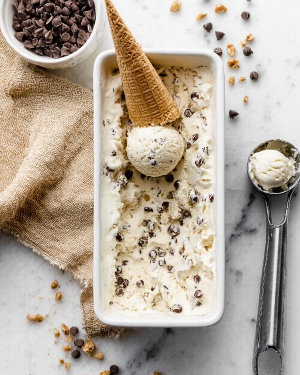 A white rectangular container is filled with creamy white ice cream mixed with chocolate chips and small nut pieces evenly spread throughout. On top, a single scoop of the ice cream holds a light brown waffle cone placed upside down. Around the container are scattered chocolate chips and a silver ice cream scoop with some ice cream residue inside. The scene is set on a white marbled surface with a beige textured cloth to the left, and a white bowl filled with chocolate chips also placed on the left side. photo taken with an iphone --ar 4:5 --v 7