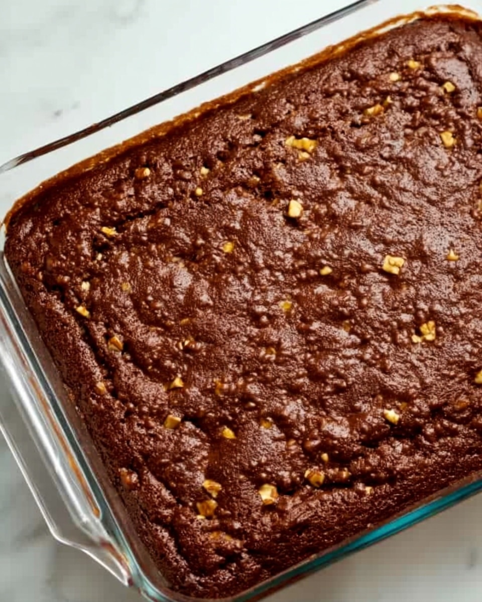 A rectangular glass dish filled with a thick, rich brown baked dessert that has a slightly uneven, bumpy surface texture. Small bits of lighter-colored nuts are scattered throughout the top layer, adding a bit of contrast. The dish is placed on a white marbled surface, and the sides of the dish show the dense, moist texture of the dessert inside. Photo taken with an iphone --ar 4:5 --v 7
