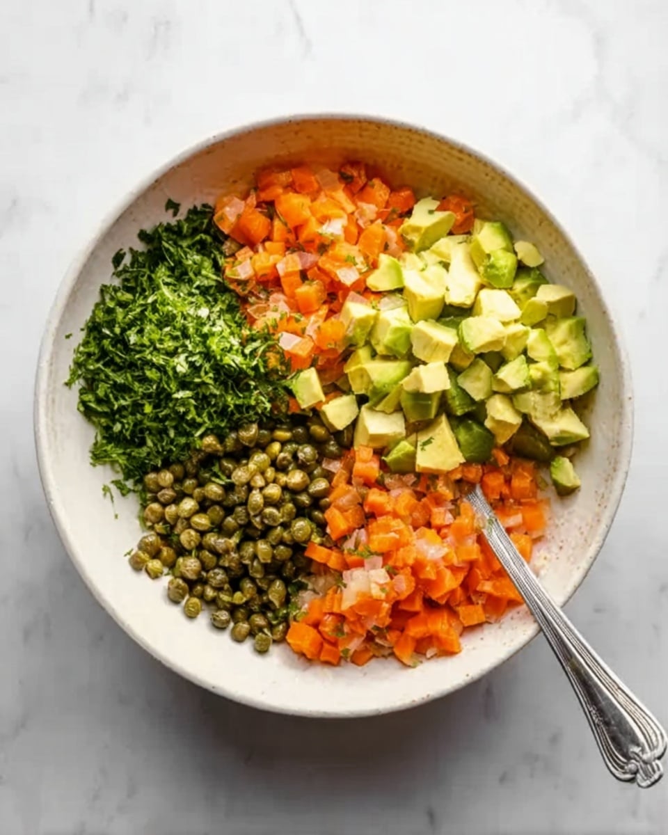 A white bowl sits on a white marbled surface, filled with four separate layers of finely chopped ingredients arranged in sections. On the top left is a pile of small green herbs, next to it on the top right are pale yellow diced pieces of avocado. Below the avocado, there are tiny orange cubes of carrot, and to the bottom left, small green capers fill that section. A silver spoon rests inside the bowl, leaning across the layers. The photo taken with an iphone --ar 4:5 --v 7
