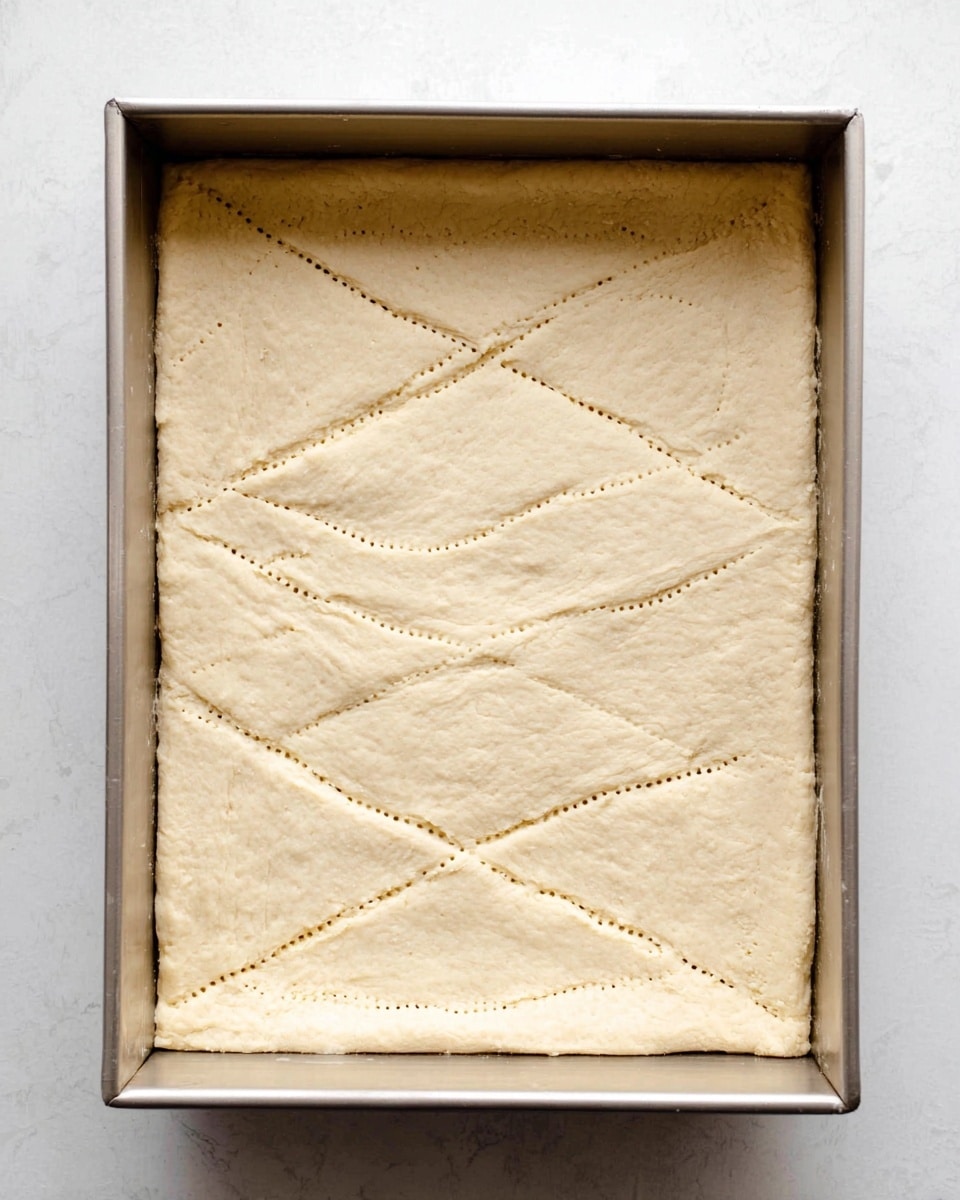 The image shows a rectangular baking pan with a raw dough layer spread evenly inside it. The dough is light beige in color and smooth in texture, with small dotted lines pressed into the surface forming a simple pattern of rectangles and diagonal lines. The edges of the dough have a slight, natural curve upward against the pan sides. The pan rests on a white marbled surface. photo taken with an iphone --ar 4:5 --v 7