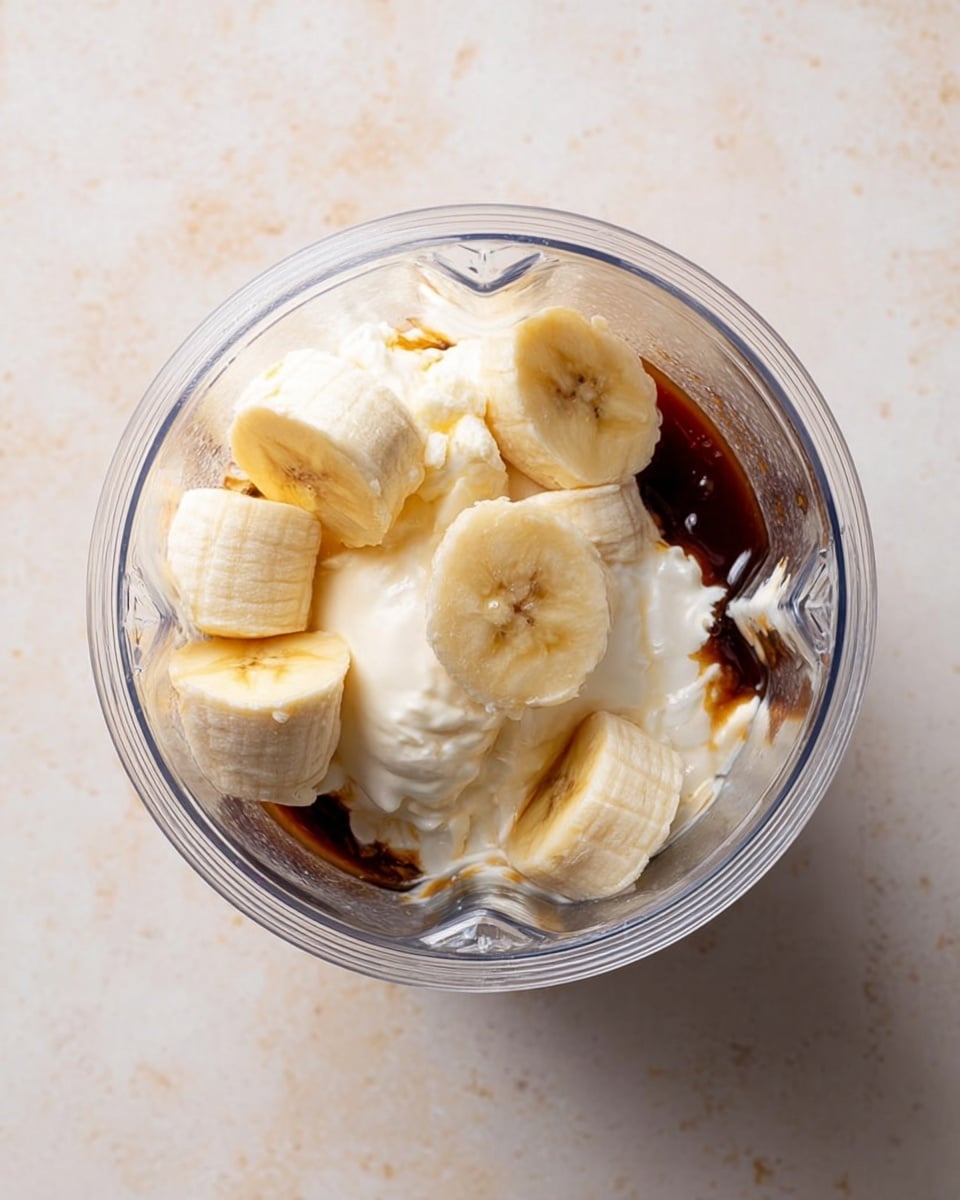 The image shows a clear blender cup filled with several layers of ingredients: at the bottom, there is a dark brown syrup-like layer, followed by a thick white cream layer on the sides, and on top, several large banana pieces in pale yellow are arranged. The blender cup is placed on a white marbled surface. The photo is taken from above with soft lighting and no shadows. Photo taken with an iphone --ar 4:5 --v 7