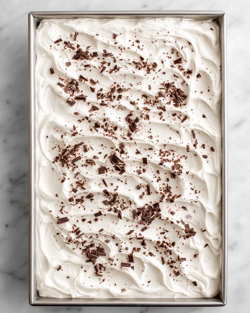 A rectangular metal pan filled with a thick layer of smooth, white whipped cream that covers the entire surface, with soft swirled patterns on top. Small, uneven pieces of dark brown chocolate shavings are sprinkled evenly across the whipped cream, adding texture and contrast. The pan sits on a white marbled surface, creating a clean and bright background. photo taken with an iphone --ar 4:5 --v 7