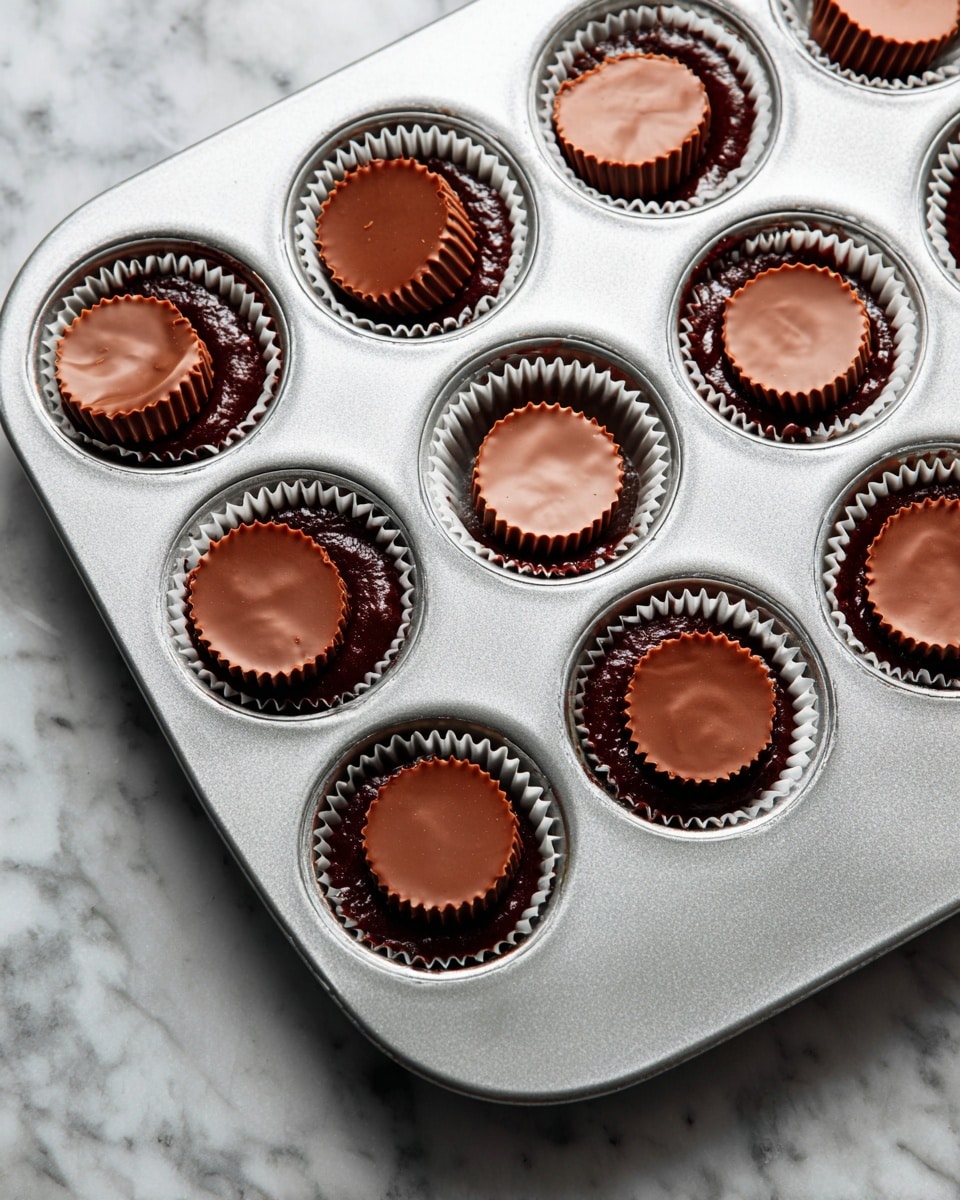 A silver muffin tray filled with dark chocolate batter in each cup, with smooth, round peanut butter cups pressed into the center of some cups. The peanut butter cups have a shiny milk chocolate appearance with ridged edges. A woman's hand with blue nail polish is pressing one peanut butter cup into the batter. The tray is on a white marbled surface. photo taken with an iphone --ar 4:5 --v 7