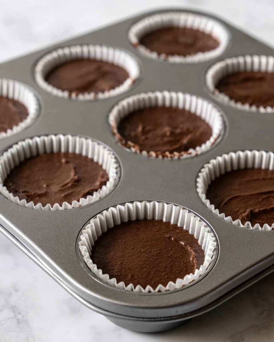 The image shows a metal cupcake tray with six cups filled with smooth, dark brown batter. Each cup is lined with a white paper cupcake liner that has a slightly crinkled edge. The batter fills each liner evenly, with a matte texture and no toppings or extra layers visible. The tray rests on a white marbled surface, and the lighting softly highlights the texture of the batter and the metal tray. photo taken with an iphone --ar 4:5 --v 7