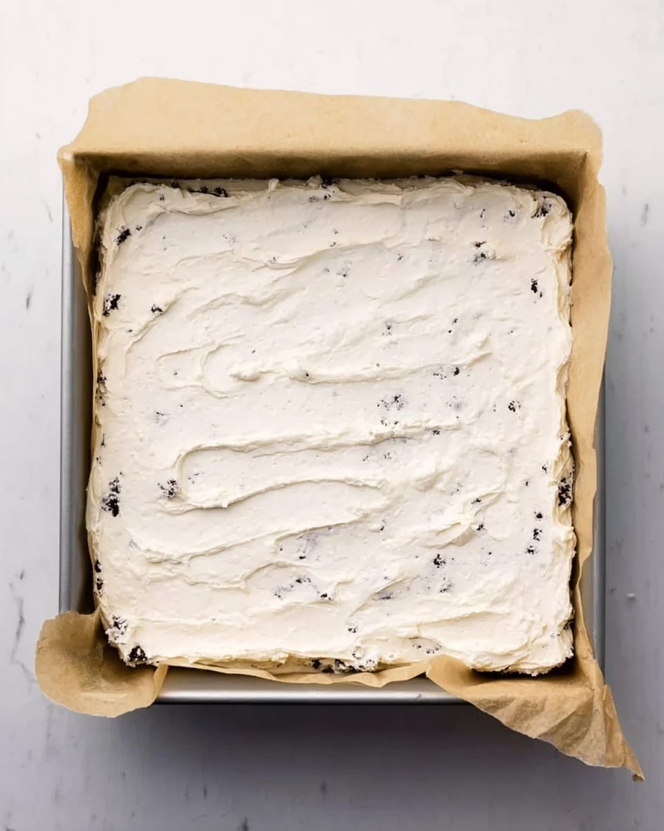 A square baking pan lined with brown parchment paper holds a single layer of thick, smooth white cream mixed with small dark cookie bits, spread evenly across the whole surface with slight texture showing some utensil marks. The pan rests on a white marbled surface. photo taken with an iphone --ar 4:5 --v 7