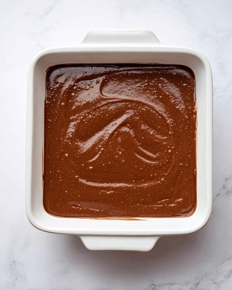 A white square ceramic baking dish with rounded handles on each side is filled with a smooth, thick chocolate batter. The batter has a shiny, rich dark brown surface with visible soft swirls and slight bubbles, evenly spread to fill the dish almost to the edges. The dish is placed on a white marbled surface, showing a clean and bright setting. Photo taken with an iphone --ar 4:5 --v 7