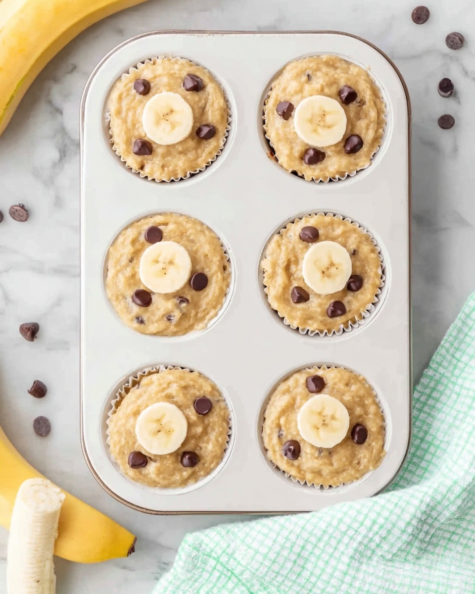 The image shows a white metal muffin tray filled with nine banana muffins batter, each topped with a slice of banana. The batter is light brown with small dark chocolate chips scattered inside. The muffin tray is placed on a white marbled surface with a light green and white checkered cloth partially visible at the bottom right corner. To the left of the tray, there is a peeled banana with one slice resting next to it and a few loose chocolate chips. The overall scene is bright and clean. Photo taken with an iphone --ar 4:5 --v 7