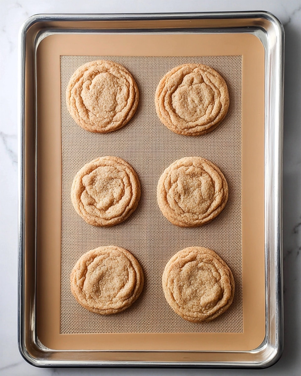 Six round cookies with a light brown color and a slightly cracked, soft texture lie evenly spaced on a tan silicone mat inside a silver baking tray. The cookies have a raised outer edge with a rough, swirled pattern in the center. The baking tray rests on a white marbled surface. Photo taken with an iphone --ar 4:5 --v 7