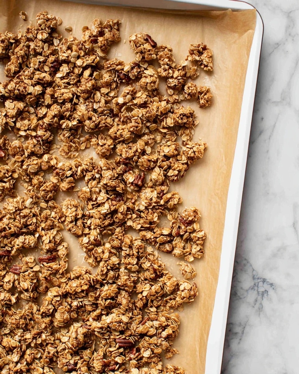 The image shows a single layer of toasted granola spread unevenly on a white baking tray lined with brown parchment paper. The granola clusters are golden brown with visible oats and pieces of nuts scattered throughout, creating a rough texture with small and large clusters. The tray edges are visible, and the background is a white marbled surface. photo taken with an iphone --ar 4:5 --v 7