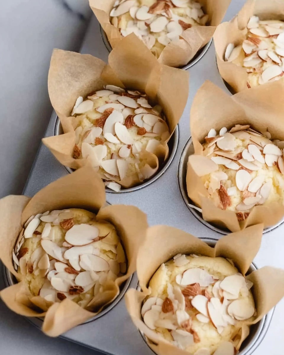 The image shows a close-up of six muffin cups lined with tan parchment paper. Each cup is filled with a pale yellow batter, and the tops are scattered with thin, irregularly shaped almond slices that have light brown edges. The muffin tray is silver and placed on a white marbled surface. The cups are evenly spaced and the parchment paper edges rise above the batter inside each cup, creating folds and peaks around the edges. Photo taken with an iphone --ar 4:5 --v 7
