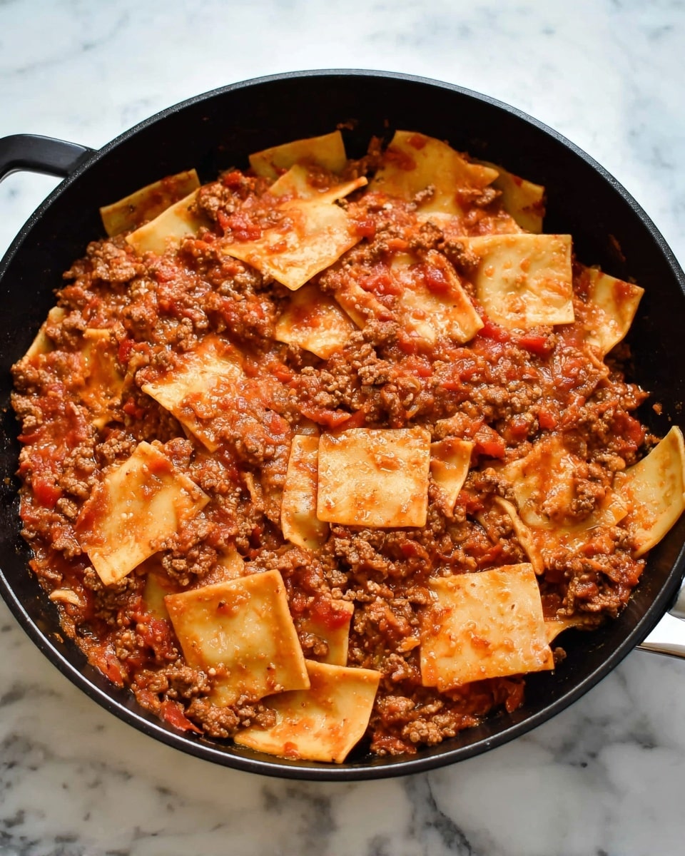 The image shows a black skillet filled with a mixture of thick, red meat sauce and flat, square pasta pieces. The sauce has a chunky texture with pieces of ground meat and small bits of tomato, giving a rich, hearty look. The pasta squares are soft, some slightly overlapping each other, coated in the sauce with a pale yellow color softened by the red sauce. The skillet sits on a surface with a white marbled texture. photo taken with an iphone --ar 4:5 --v 7