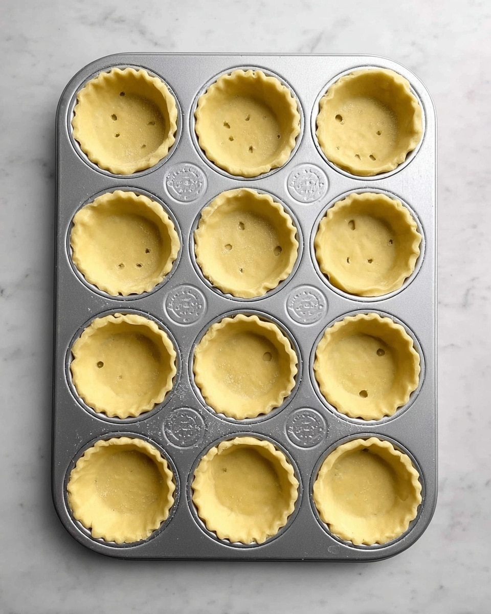 A metal muffin tray with twelve round cups holds raw tart shells made from light yellow dough. Each tart shell lining the cups has slightly uneven edges, and small fork holes are visible in the bottom center of each shell, arranged in a grid shape. The tray is placed on a white marbled surface, showing a clean, simple setting. The photo is taken from above, capturing all tart shells clearly in even light, photo taken with an iphone --ar 4:5 --v 7