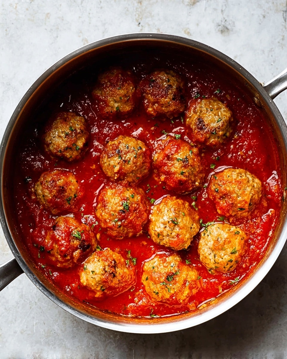A pot filled with shiny meatballs sitting in a thick red tomato sauce, the meatballs are light brown with herbs visible inside them, clustered closely together and covered partially by the sauce, the pot is silver on the outside and rests on a white marbled surface, the sauce looks smooth and rich with chunks of tomato visible, the lighting highlights the texture of the meatballs and the glossy sauce, photo taken with an iphone --ar 4:5 --v 7