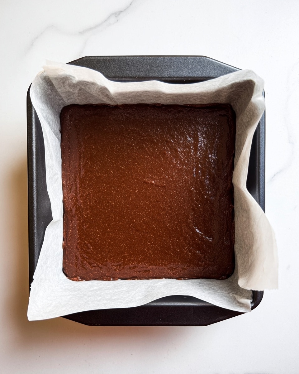 A square black baking pan lined with white parchment paper holds a smooth, even layer of dark brown chocolate batter spread out to the edges. The parchment paper edges are wrinkled and rise slightly above the batter. The pan is placed on a white marbled surface. photo taken with an iphone --ar 4:5 --v 7