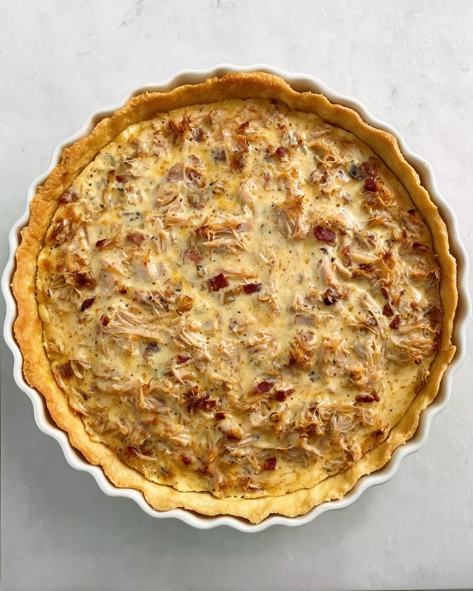 The image shows a round tart with a golden-brown crust that has a scalloped edge. The filling is creamy and light brown with visible pieces of shredded ingredients, possibly chicken and bits of bacon, mixed throughout. The surface of the tart has a slightly uneven texture from the shredded pieces inside. The tart is inside a white ceramic dish, placed on a white marbled surface. photo taken with an iphone --ar 4:5 --v 7