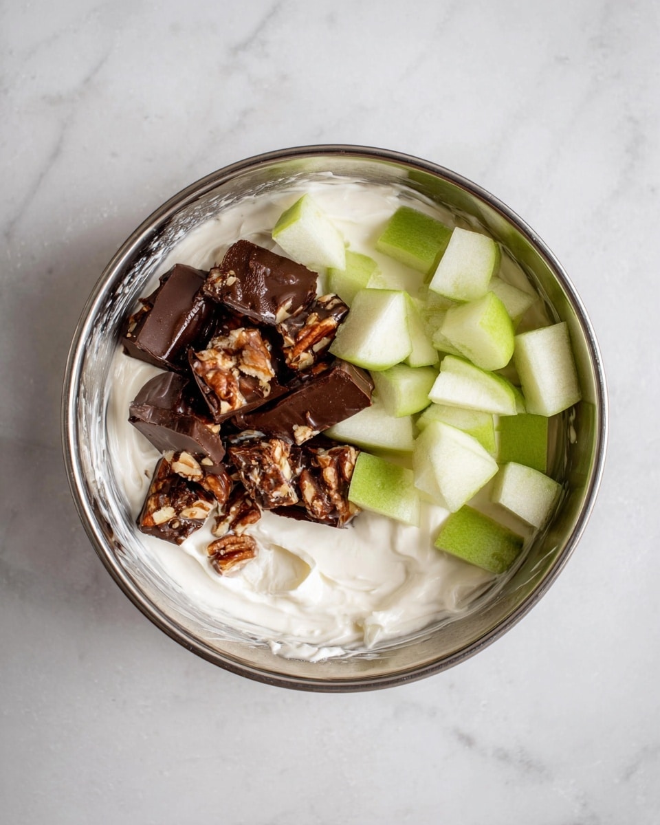 A shiny silver bowl holds a thick white creamy base that fills most of the bowl. On top, there are small green apple pieces with smooth edges on the right side, and chunky chocolate-covered nut bars with dark brown glossy chocolate and visible nuts stacked on the left side. The inside edge of the bowl has some of the white cream sticking to it. The bowl sits on a white marbled surface. photo taken with an iphone --ar 4:5 --v 7