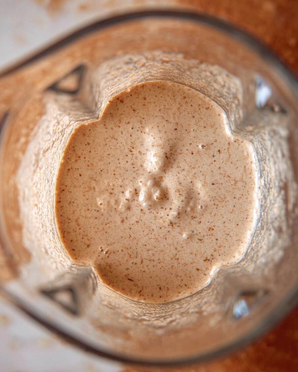 A close-up view from above of a clear blender filled with a smooth light brown liquid mixture that has small dark specks evenly spread throughout. The blender is partially transparent with reflections and some texture details visible. The background is a white marbled surface. Photo taken with an iphone --ar 4:5 --v 7