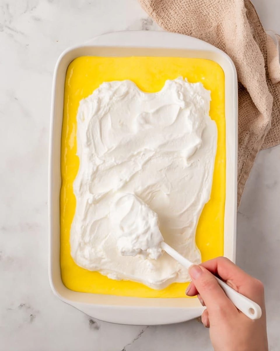 The image shows a two-layer dessert in a white rectangular baking dish placed on a white marbled surface. The bottom layer is smooth and bright yellow, covering the entire base of the dish. Above it, a thick and fluffy white cream layer is being spread evenly with a white spoon held by a woman's hand coming from the bottom right corner. A soft beige cloth is slightly visible in the upper right background. Photo taken with an iphone --ar 4:5 --v 7