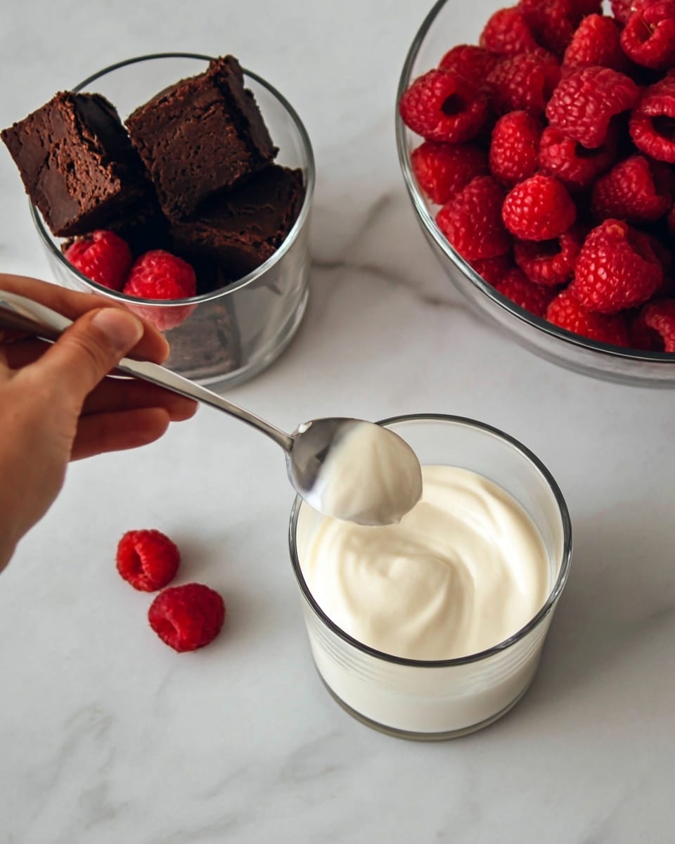 The image shows a clear glass cup with a creamy white layer filling most of it; a woman's hand holds a spoon that is lifting some creamy white mixture from the cup. Next to it, there is another clear glass cup filled with the same creamy white mix, and above that, a clear glass bowl is full of bright red raspberries. To the left, a clear glass cup contains pieces of dark brown brownie and a few raspberries on top and around the brownies. All items rest on a white marbled texture surface. Photo taken with an iphone --ar 4:5 --v 7