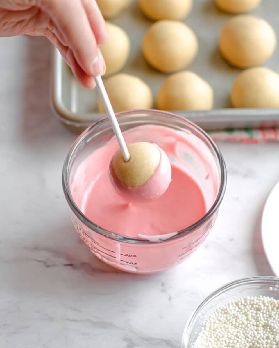 A close-up shot shows a clear glass measuring cup filled with smooth, light pink icing. A pale beige round dough ball is being dipped into the icing with a white stick, held by a woman's hand visible on the left side. In the top part of the image, there is a metal tray with several similar round dough balls, all light beige in color and smooth textured. The background is a white marbled surface with a softly blurred white dish partly visible on the right side and a clear container filled with small white sprinkles near the bottom center. Photo taken with an iphone --ar 4:5 --v 7