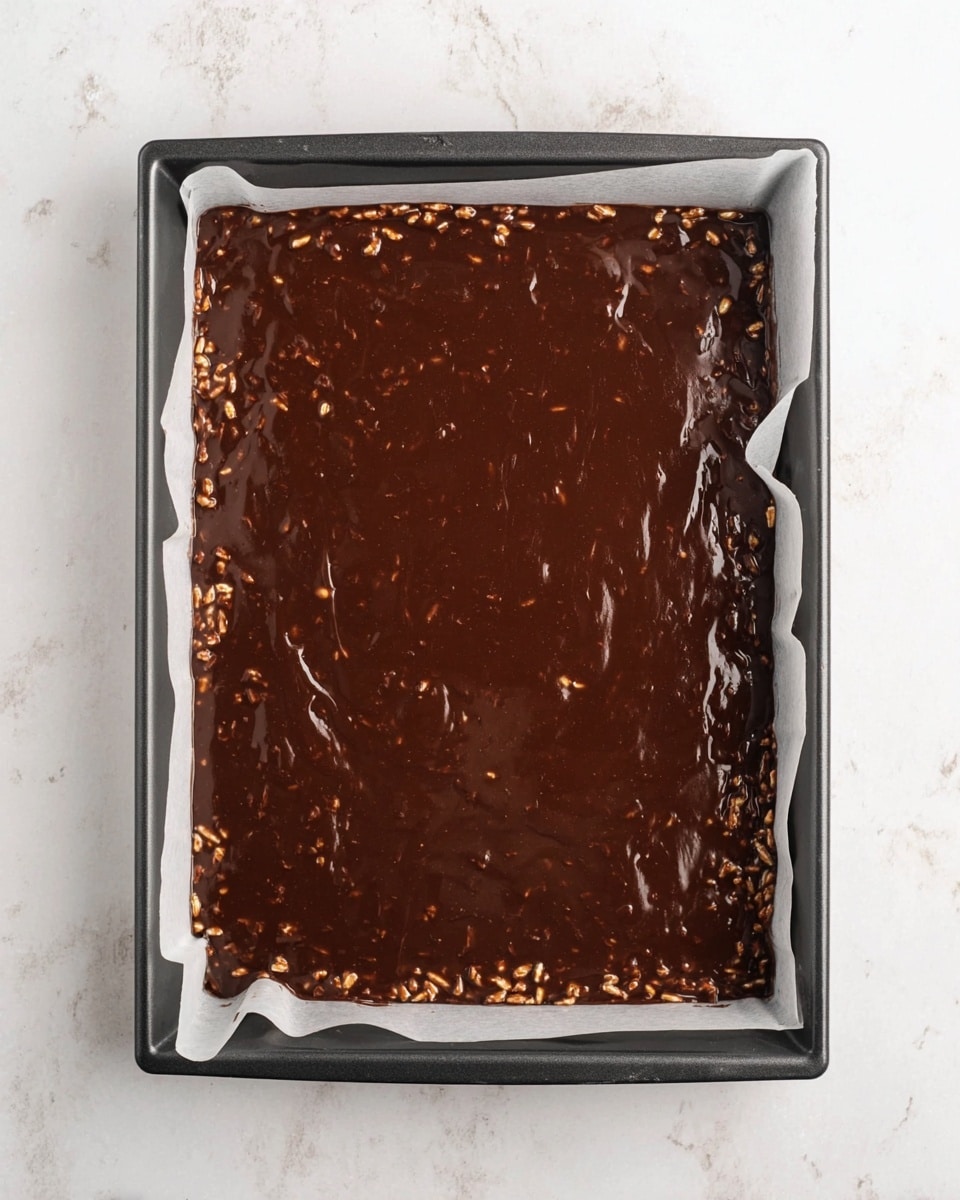 A rectangular dark baking tray lined with white paper holds a thick layer of dark brown chocolate mixture with small pieces of nuts or crisped rice visible throughout. The glossy chocolate surface looks smooth but uneven, showing the texture of the ingredients mixed inside. The tray sits on a white marbled surface, giving a clean and bright background. photo taken with an iphone --ar 4:5 --v 7
