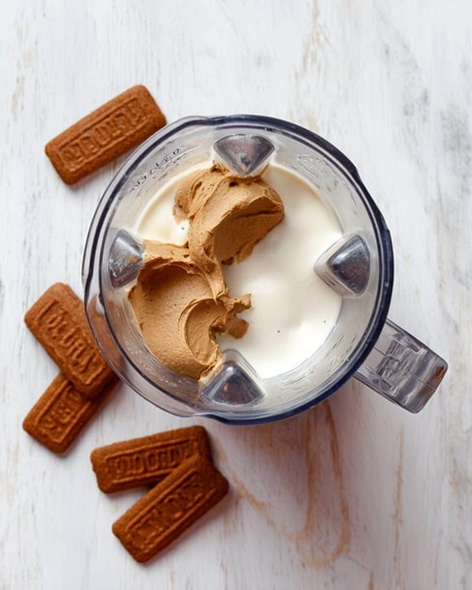 The image shows the top view of a blender with two main layers inside: at the bottom, a smooth white creamy layer, and on top of it, two dollops of thick light brown spread. Outside the blender, four brown rectangular biscuits with text on them are placed on a white marbled surface, arranged around the blender. Photo taken with an iphone --ar 4:5 --v 7