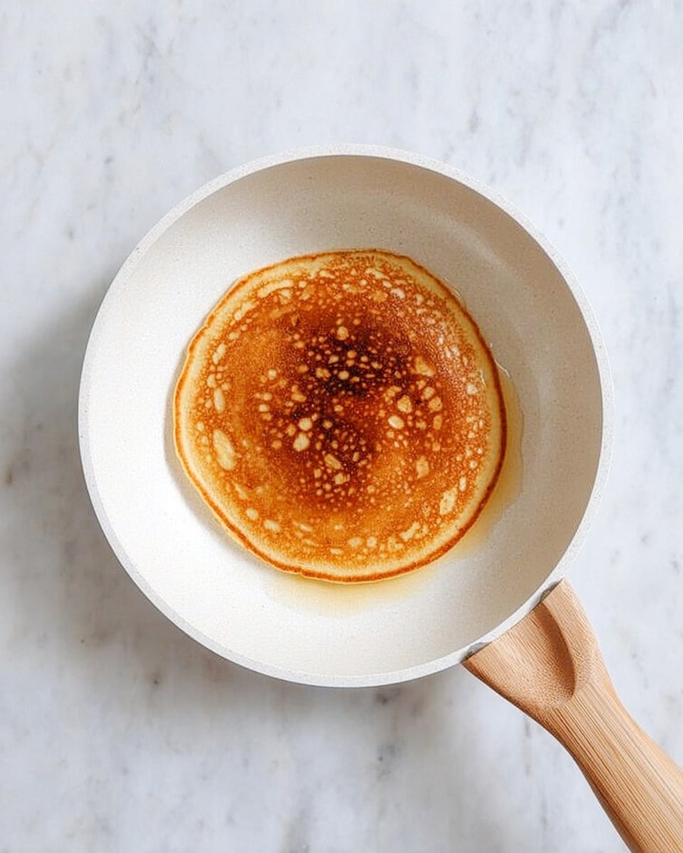 A single round pancake with a browned top and lighter center sits in the middle of a white frying pan with a light wood handle. The pancake has uneven golden brown edges and a soft texture. The background is a white marbled surface. photo taken with an iphone --ar 4:5 --v 7