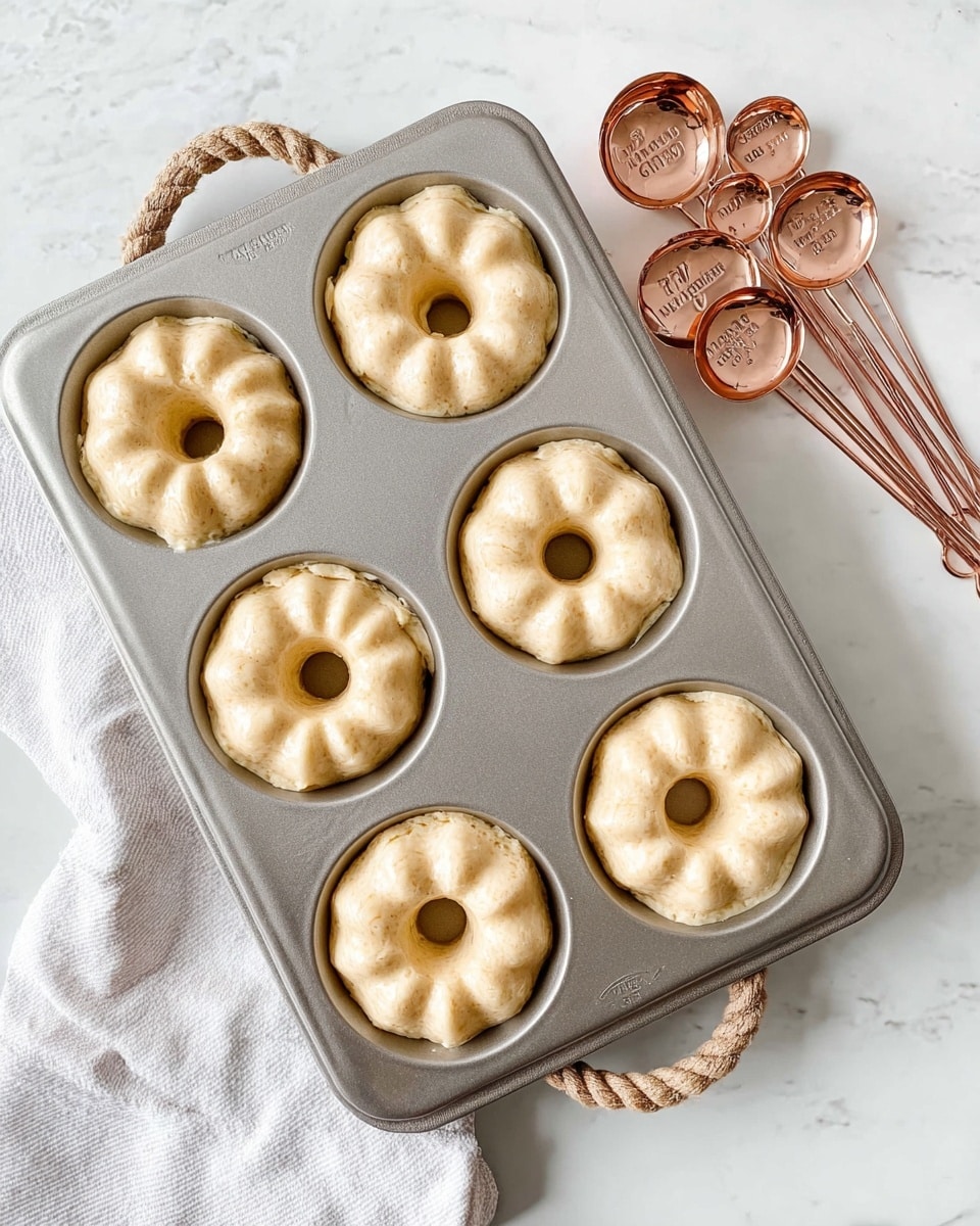 A metallic baking pan with six small bundt cake molds is filled with smooth, light beige batter that looks thick and creamy, each mold showing the batter evenly spread and slightly rounded at the top with a central hole shape visible; the pan has a textured handle with a rope design on the top left and bottom right corners. Next to the pan, a set of rose gold measuring spoons with size labels sits flat on a white marbled surface, reflecting soft light. A white cloth is partly visible on the bottom left side. The scene is bright and clean, showing the batter ready for baking. photo taken with an iphone --ar 4:5 --v 7