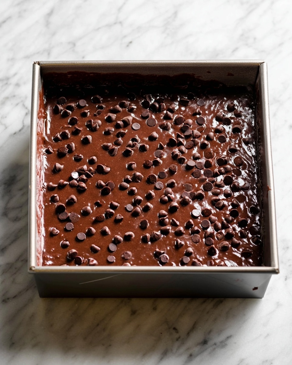 A square metal baking pan filled with a thick layer of smooth dark brown chocolate batter, sprinkled evenly with shiny chocolate chips of different sizes on top. The pan is placed on a surface with a white marbled texture, and the light reflects slightly on the wet batter, showing a glossy finish. Photo taken with an iphone --ar 4:5 --v 7
