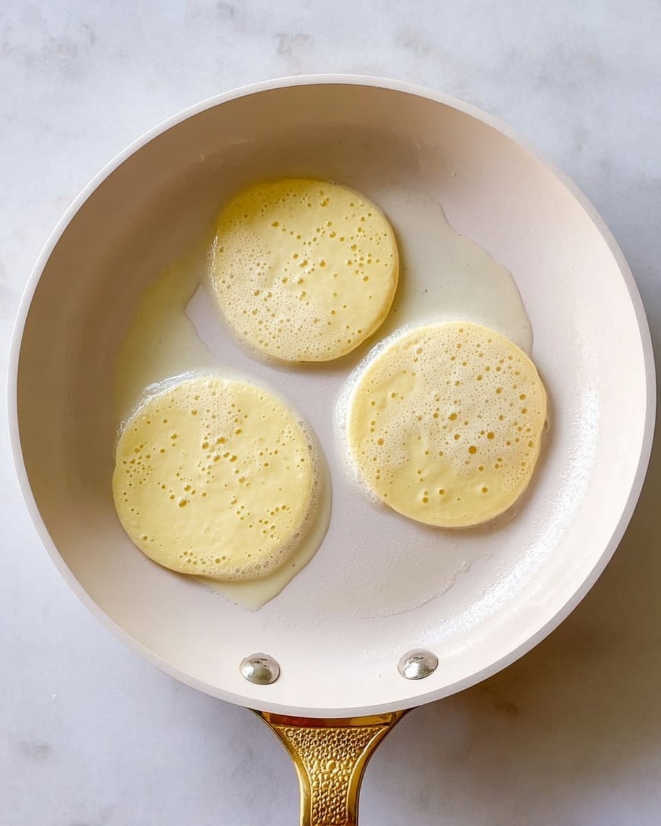 Three small light yellow pancake batter rounds cooking in a white pan with a golden handle, bubbles forming on the surface of each pancake. The pan is set on a white marbled texture background. Photo taken with an iphone --ar 4:5 --v 7
