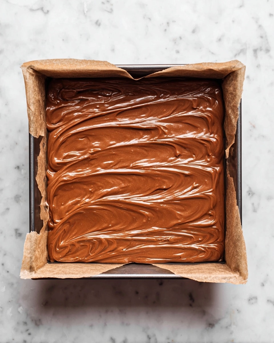 A square metal pan lined with parchment paper holds one thick, smooth layer of shiny light brown chocolate batter evenly spread inside; the batter has soft swirls and waves on its surface creating a textured look. The pan is placed on a white marbled surface. photo taken with an iphone --ar 4:5 --v 7