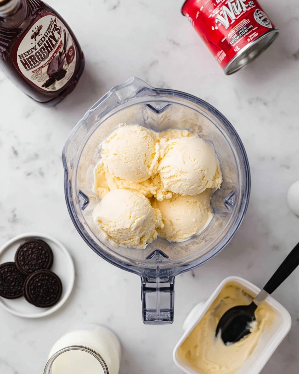 A clear blender container filled with several large scoops of pale yellow ice cream sits centered on a white marbled surface. To the top left, there is a bottle of Hershey's chocolate syrup and a red and white can of Reddi-wip. On the right side, there is a white tub of ice cream with a black spoon inside it. At the bottom left corner, a small white dish contains three dark chocolate sandwich cookies. Nearby, a small glass bottle holds white milk. The scene is bright and clean, with a simple arrangement of ingredients around the blender. Photo taken with an iphone --ar 4:5 --v 7