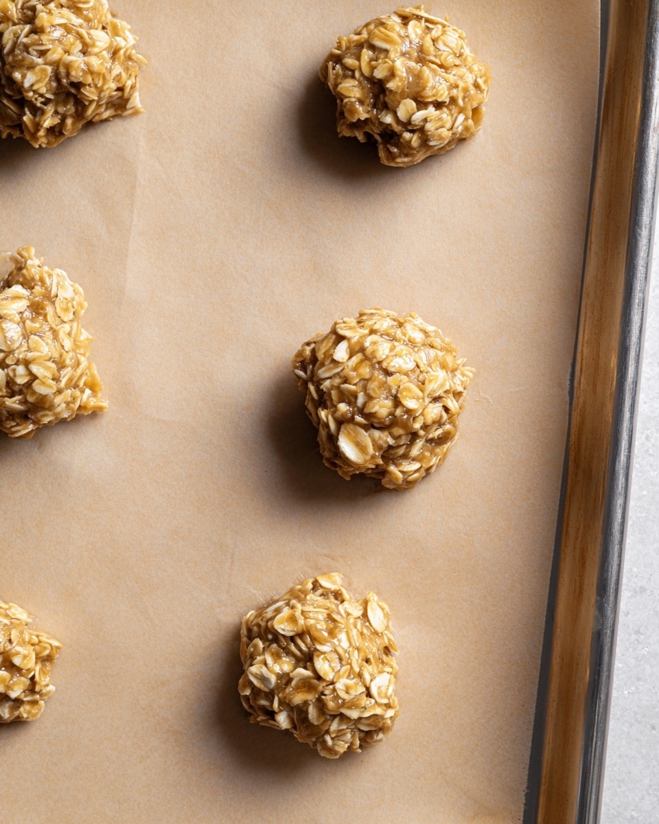 The image shows five small, round clusters of raw oatmeal dough placed unevenly on a light brown parchment paper that covers a metal baking tray. Each cluster is made up of thick rolled oats mixed with a shiny, sticky syrup, giving the dough a rough texture with visible oat pieces. The clusters are spaced apart over the parchment, which has a natural matte texture, and the baking tray’s silver edge is visible on the right. The overall look is simple and rustic with a focus on the coarse oat texture photo taken with an iphone --ar 4:5 --v 7