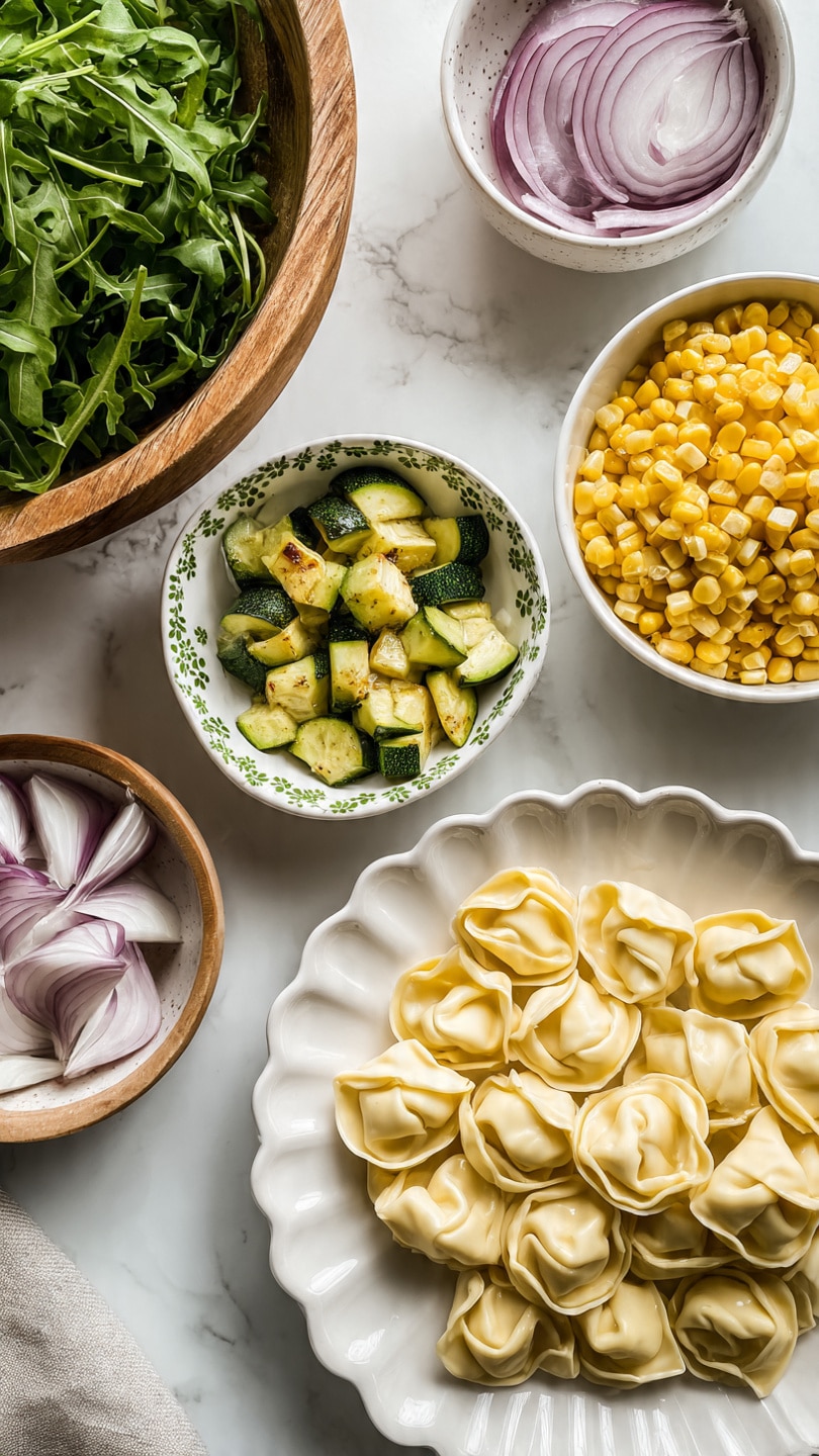 The image shows a white scalloped plate full of pale yellow tortellini pasta lined up close to each other, positioned at the bottom center. Above it, a small white bowl with green decorative spots contains chopped cooked zucchini pieces that are green with browned edges. To the top right, there is a white bowl filled with bright yellow corn kernels. Below it, a small white bowl with a brown rim contains thinly sliced light purple onions. On the left side, a large wooden bowl is filled with fresh dark green arugula leaves. All items are placed on a white marbled surface. Photo taken with an iphone --ar 4:5 --v 7