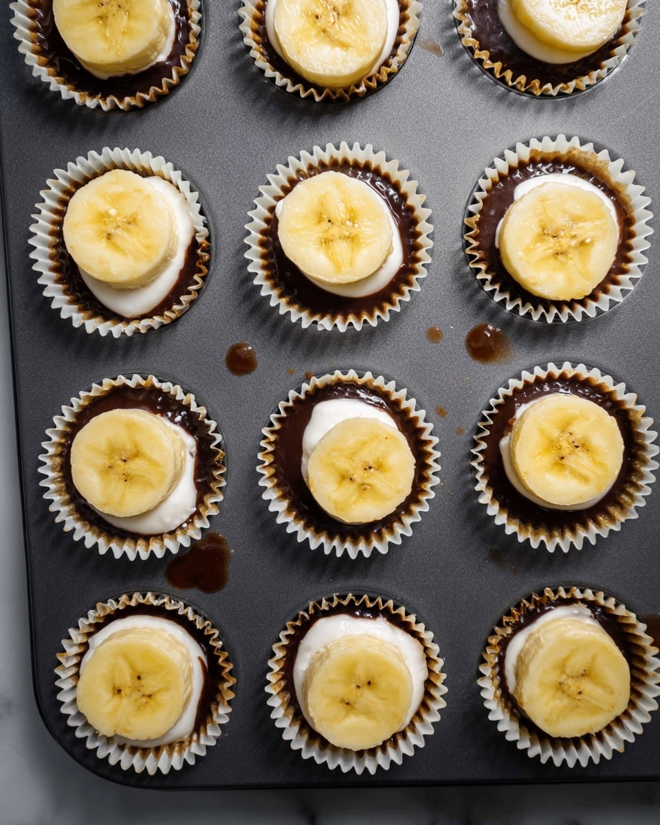 A dark gray baking tray holds 16 small white paper cupcake liners, each filled with three layers: a glossy dark brown chocolate base, a middle layer of smooth white cream, and a top layer of a round yellow banana slice with a slightly shiny surface. The banana slices have a soft texture and faint brown spots. Small droplets of chocolate are visible on the tray. The scene is set on a white marbled surface. Photo taken with an iphone --ar 4:5 --v 7