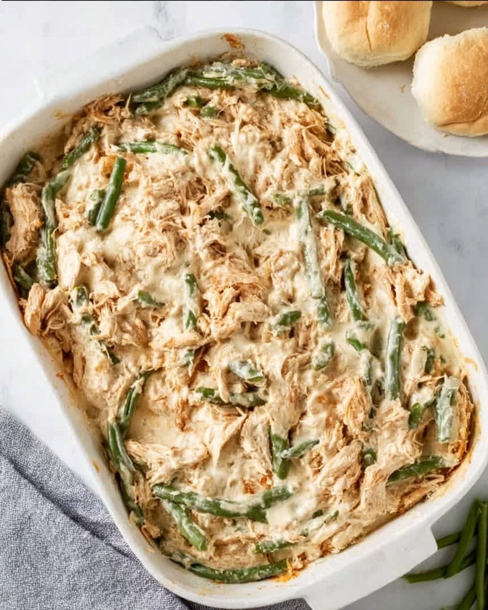 The image shows a rectangular white dish filled with a creamy baked mixture that has visible chunks of light brown and shredded pieces mixed with vibrant green beans. The texture looks soft and rich with a slightly uneven surface from the green beans scattered throughout. The dish rests on a white marbled surface, with parts of a white plate holding soft beige rolls seen in the top right corner, and a gray cloth partially under the dish at the bottom left. The overall colors are light with natural tones of beige, green, and creamy white photo taken with an iphone --ar 4:5 --v 7