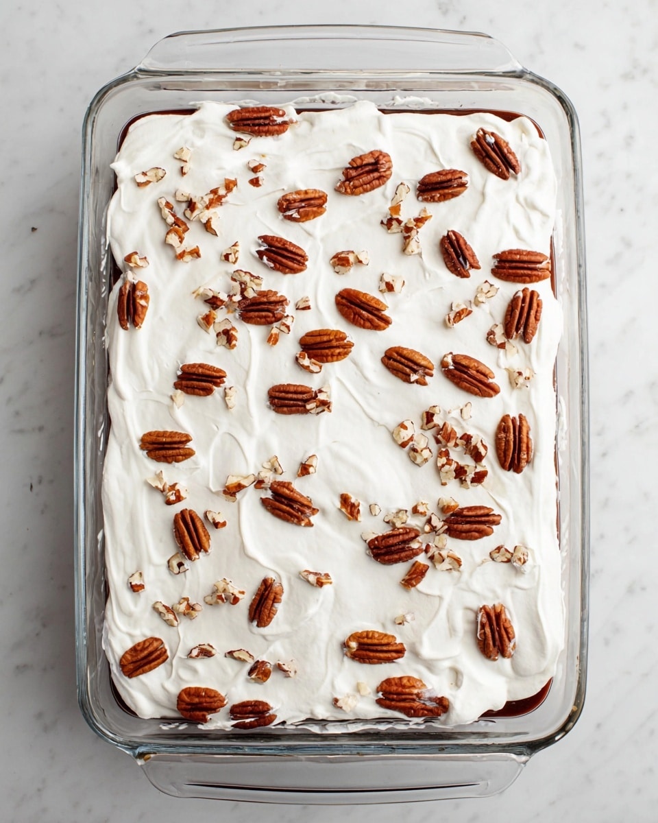 A clear rectangular glass dish filled with two visible layers: the bottom layer is dark brown and smooth, likely a cake or pudding layer, and the top layer is a thick, even spread of white whipped cream with a soft texture. Scattered evenly on top are whole and halved pecans, toasted brown with a slight shine and rough texture. The dish is placed on a white marbled surface. Photo taken with an iphone --ar 4:5 --v 7
