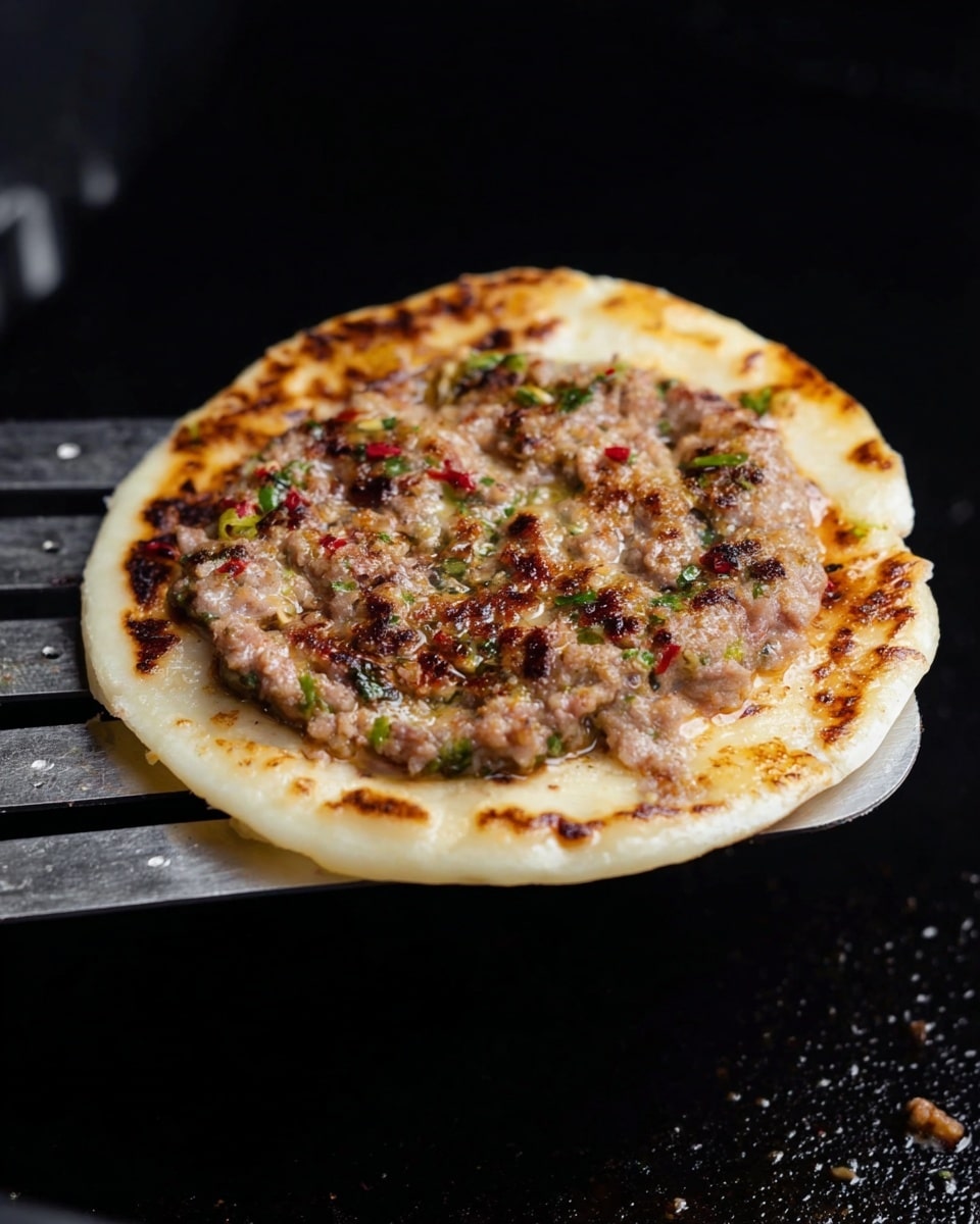 A small round flatbread is topped with a layer of cooked meat mixed with small green and red bits, showing a slightly browned and textured surface. The flatbread underneath has light golden brown spots from cooking and appears soft yet slightly crispy at the edges. The dish is held on a metal spatula over a dark cooking surface with a black background. The flatbread and meat layer contrast well with the dark background, showing a warm and meaty texture on top of the pale bread. photo taken with an iphone --ar 4:5 --v 7