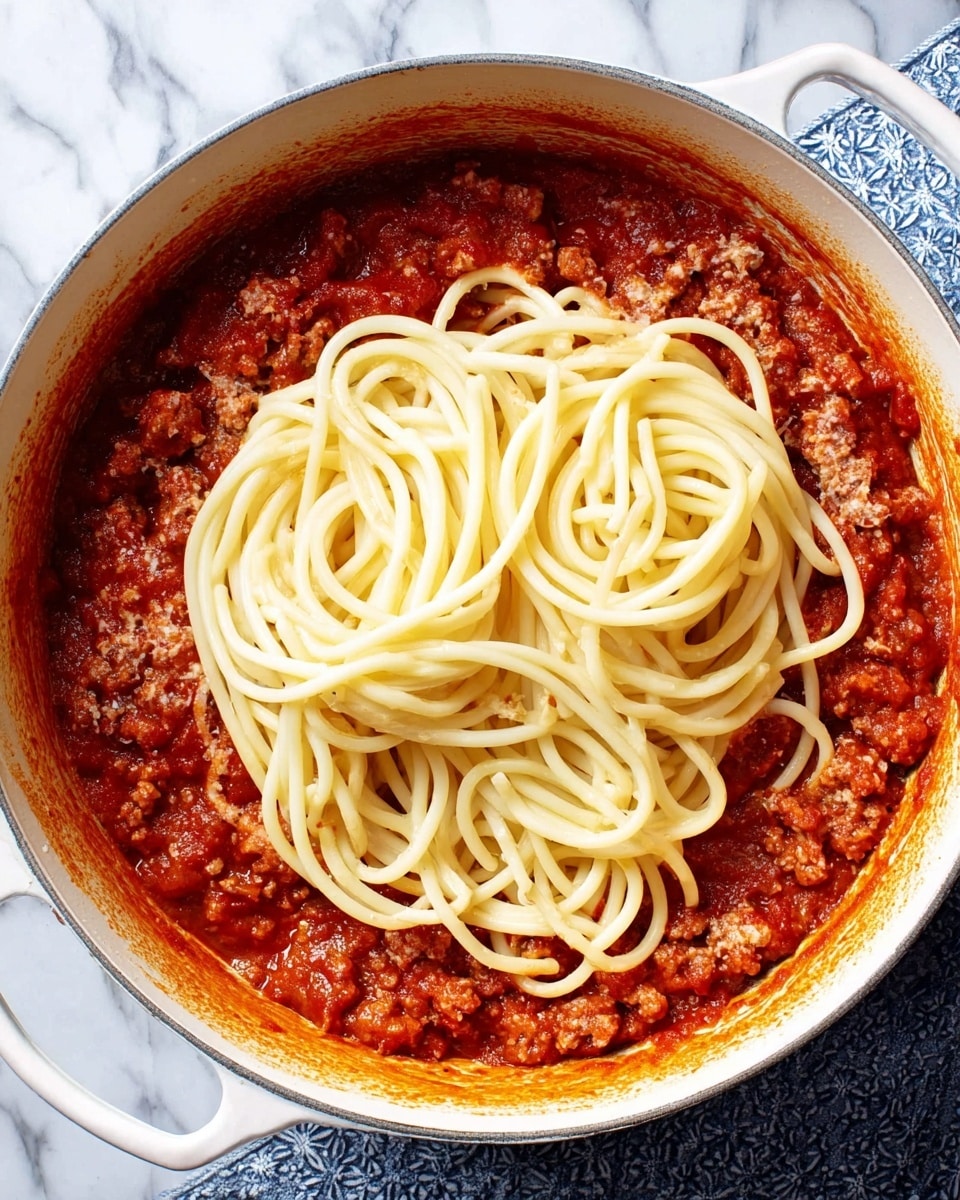The image shows a white pot filled with a thick red sauce mixed with small meat pieces, covering the bottom and sides of the pot. On top of the sauce, there is a layer of long, pale yellow pasta arranged in loose loops, piled in the center. The pot rests on a white marbled surface with a blue patterned cloth partly visible on the right side. Photo taken with an iphone --ar 4:5 --v 7