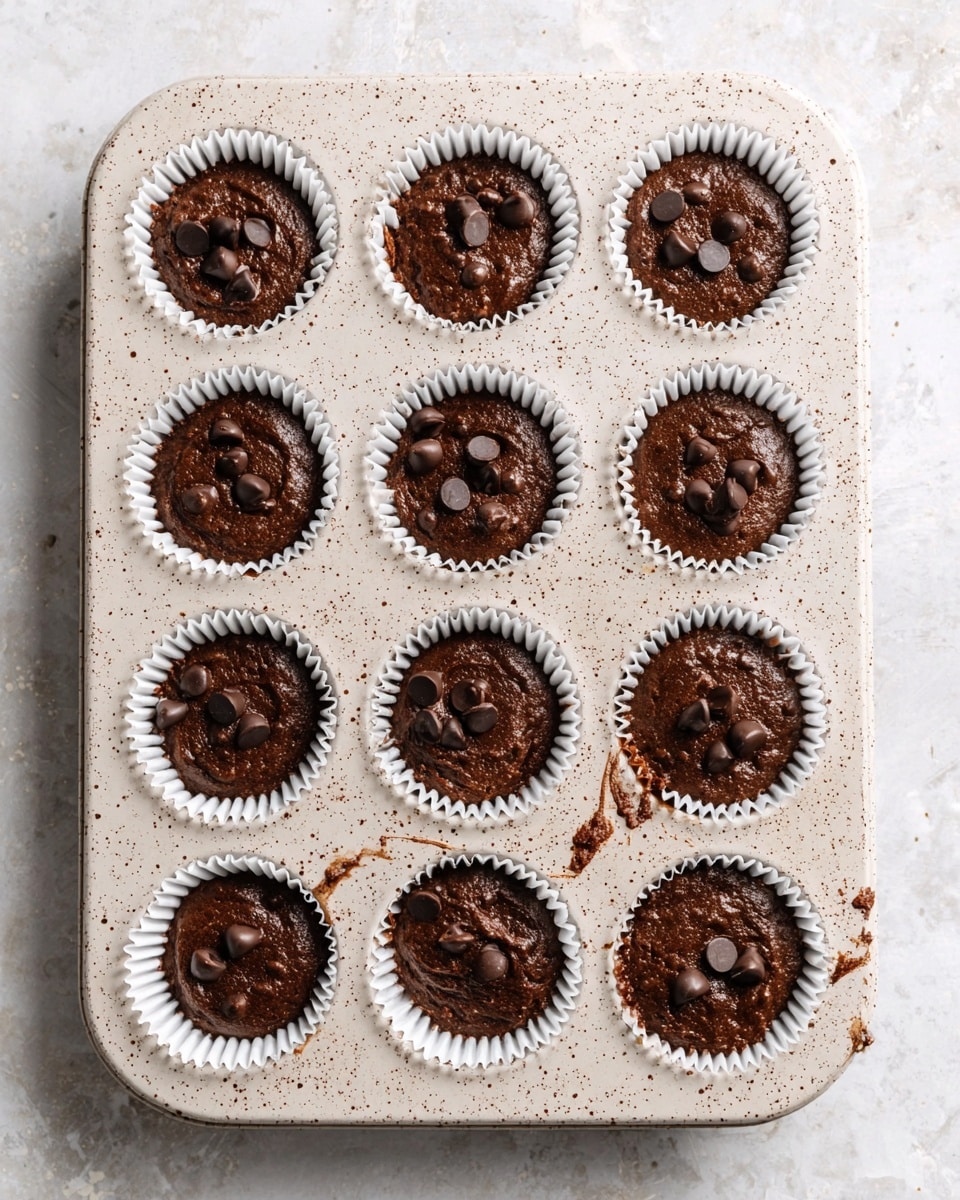 A white speckled muffin tray holds twelve white paper cupcake liners filled almost to the top with dark brown chocolate batter. Each cupcake surface is slightly uneven with small clusters of dark chocolate chips scattered on top. The tray sits on a white marbled surface with some smudges of batter visible around the edges of the liners and tray. The lighting is soft and natural, showing the rich texture of the batter clearly photo taken with an iphone --ar 4:5 --v 7