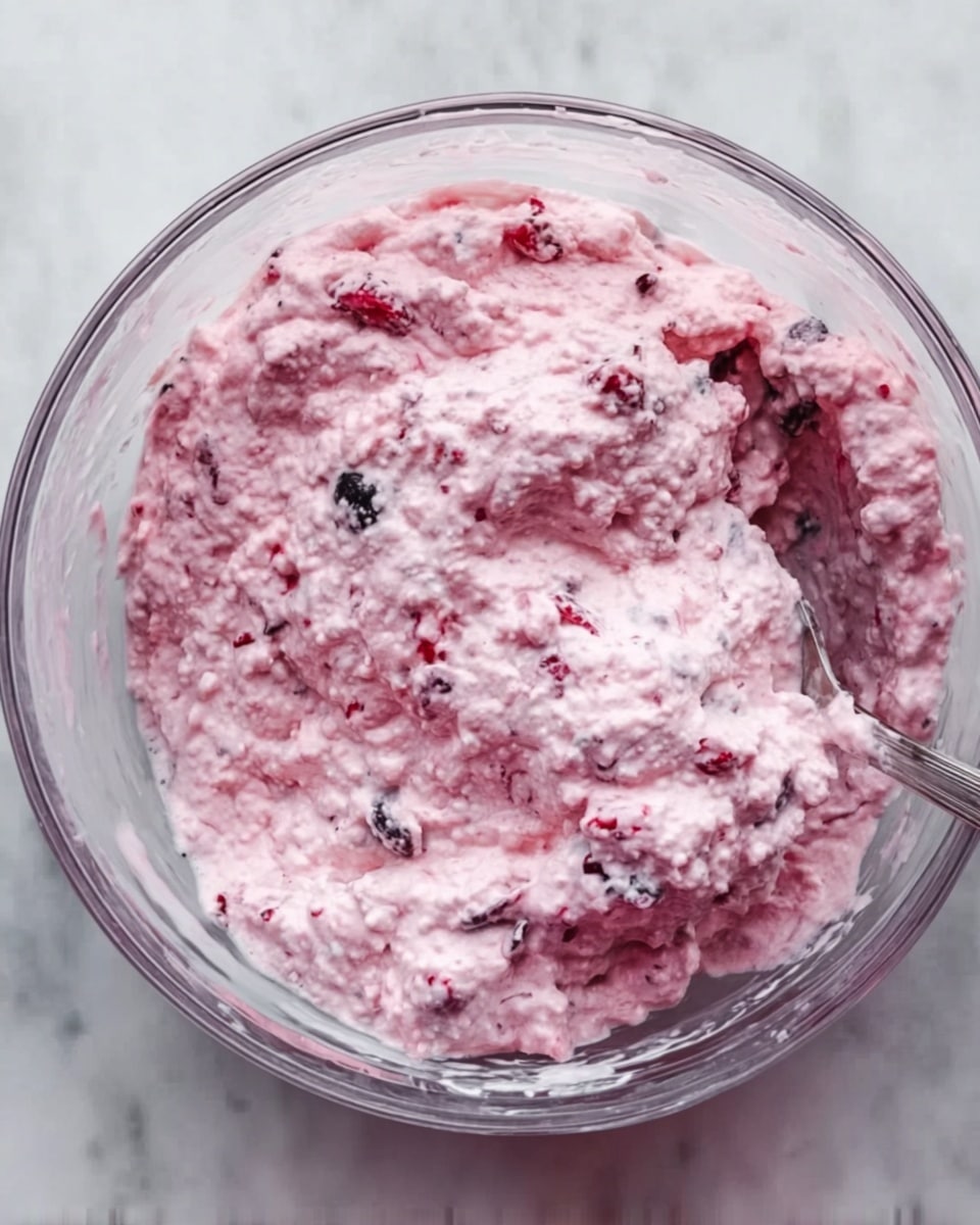 The image shows a clear glass bowl filled with a thick, creamy pink mixture that has a bumpy texture. Inside the mixture, there are small bits of red and black pieces scattered evenly through the pink base. A silver spoon is partially visible, resting on the side of the bowl, slightly covered by the mixture. The bowl is set on a white marbled surface. Photo taken with an iphone --ar 4:5 --v 7