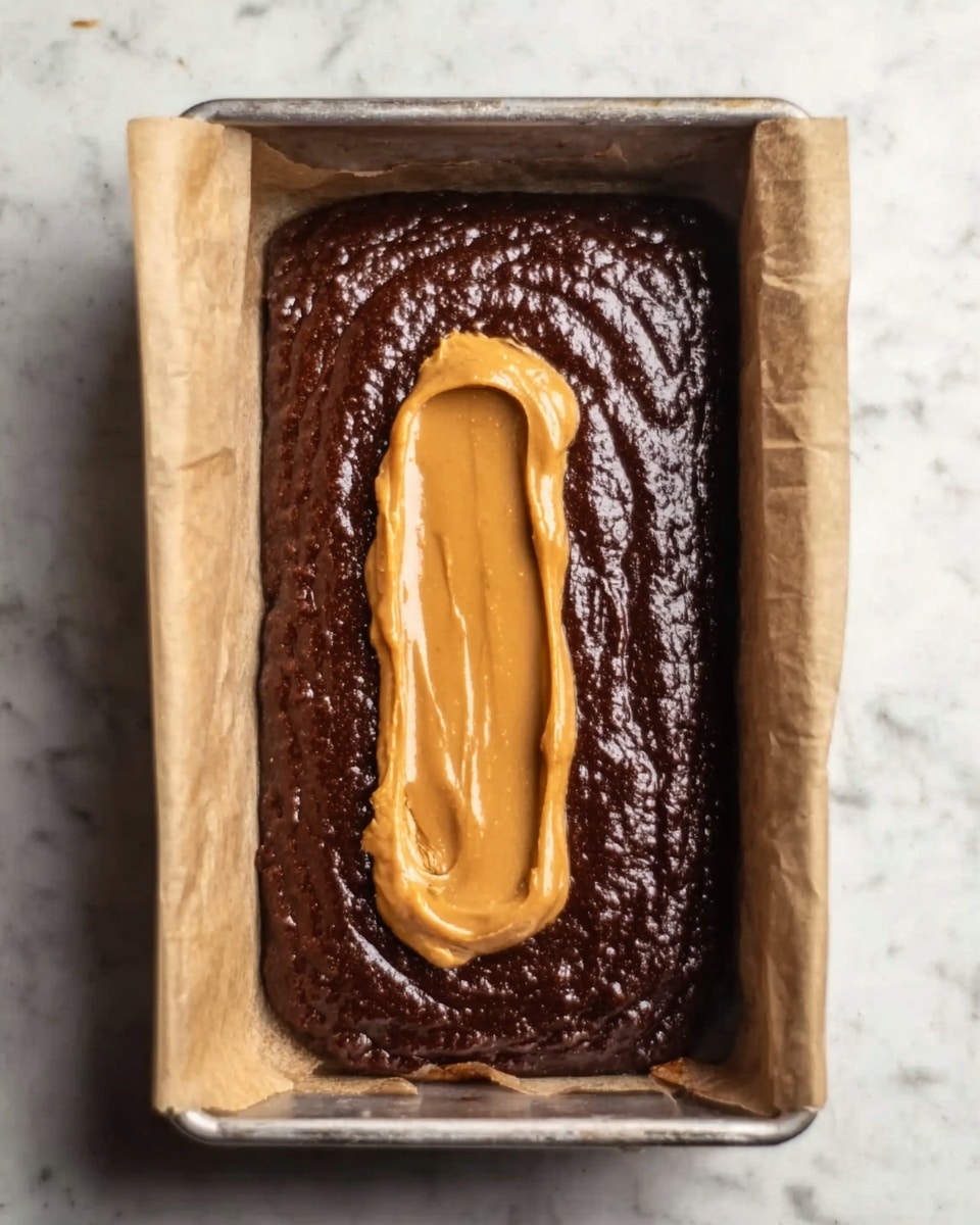 The image shows a baking tray lined with parchment paper containing two layers of dessert batter. The bottom layer is a thick, rich dark brown chocolate batter filling the entire tray. On top of this layer, in the center, is a smaller elongated dollop of smooth, caramel-colored peanut butter batter with a creamy texture, slightly spread but not covering the chocolate layer fully. The baking tray sits on a white marbled surface. Photo taken with an iphone --ar 4:5 --v 7
