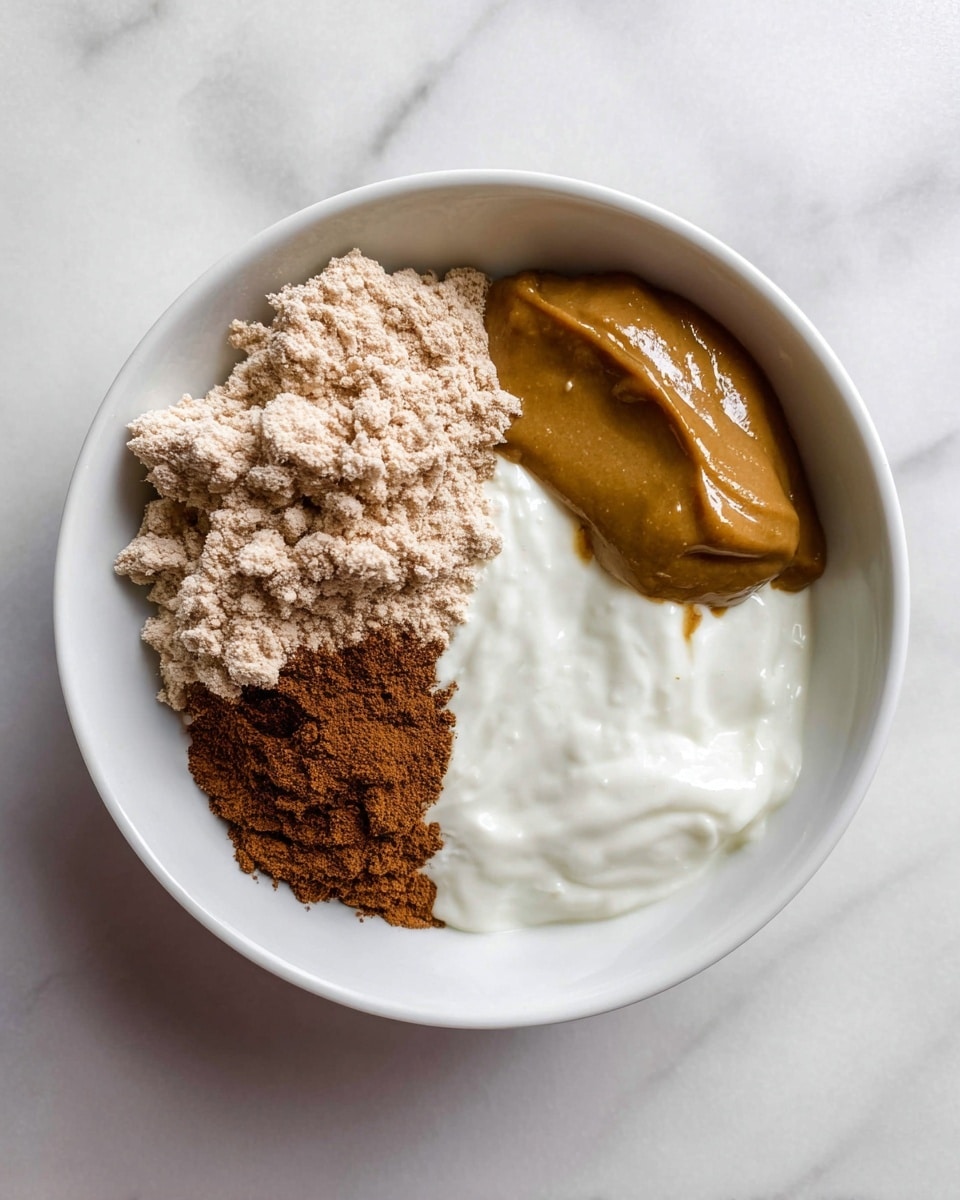 A white bowl on a white marbled surface holds four distinct layers of ingredients that are not mixed. The largest portion in the bowl is a smooth and thick white creamy layer at the bottom right. To its top right, there is a rich golden brown dollop with a shiny texture, sitting neatly above the white layer. On the left side of the bowl, a clumpy light brown powder forms a rough pile, occupying almost half of the bowl’s space. Between the white creamy layer and the light brown powder, a thin layer of darker brown fine powder spices the mix, adding a contrast in color and texture. photo taken with an iphone --ar 4:5 --v 7